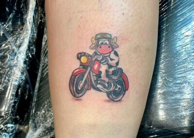 A tattoo of a cartoon Cow riding on a Motorbike on the back of the calf