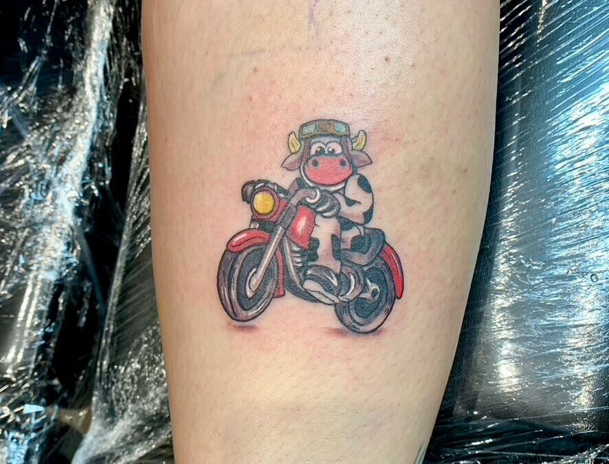 A tattoo of a cartoon Cow riding on a Motorbike on the back of the calf